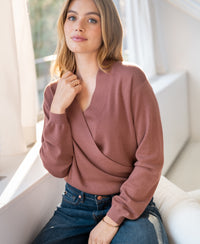 A woman wears a PLEIN PUBLIQUE wrap sweater in desert rose near a sunlit window.