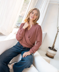 Woman wearing a desert rose wrap sweater sits thoughtfully on a white couch by the window.