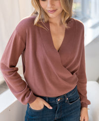 Woman wearing a desert rose wrap sweater by PLEIN PUBLIQUE, indoors with a hand in her pocket.