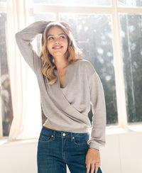 Woman smiling in sunlight, wearing a beige sand melange wrap sweater by large windows.