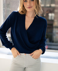 Navy blue crossover sweater by PLEIN PUBLIQUE shown on a model standing by a sunlit window, hands in pockets.