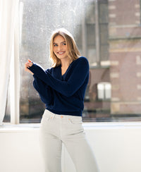 Woman wearing PLEIN PUBLIQUE's LES CINQ Navy Wrap Sweater stands by a sunny window.