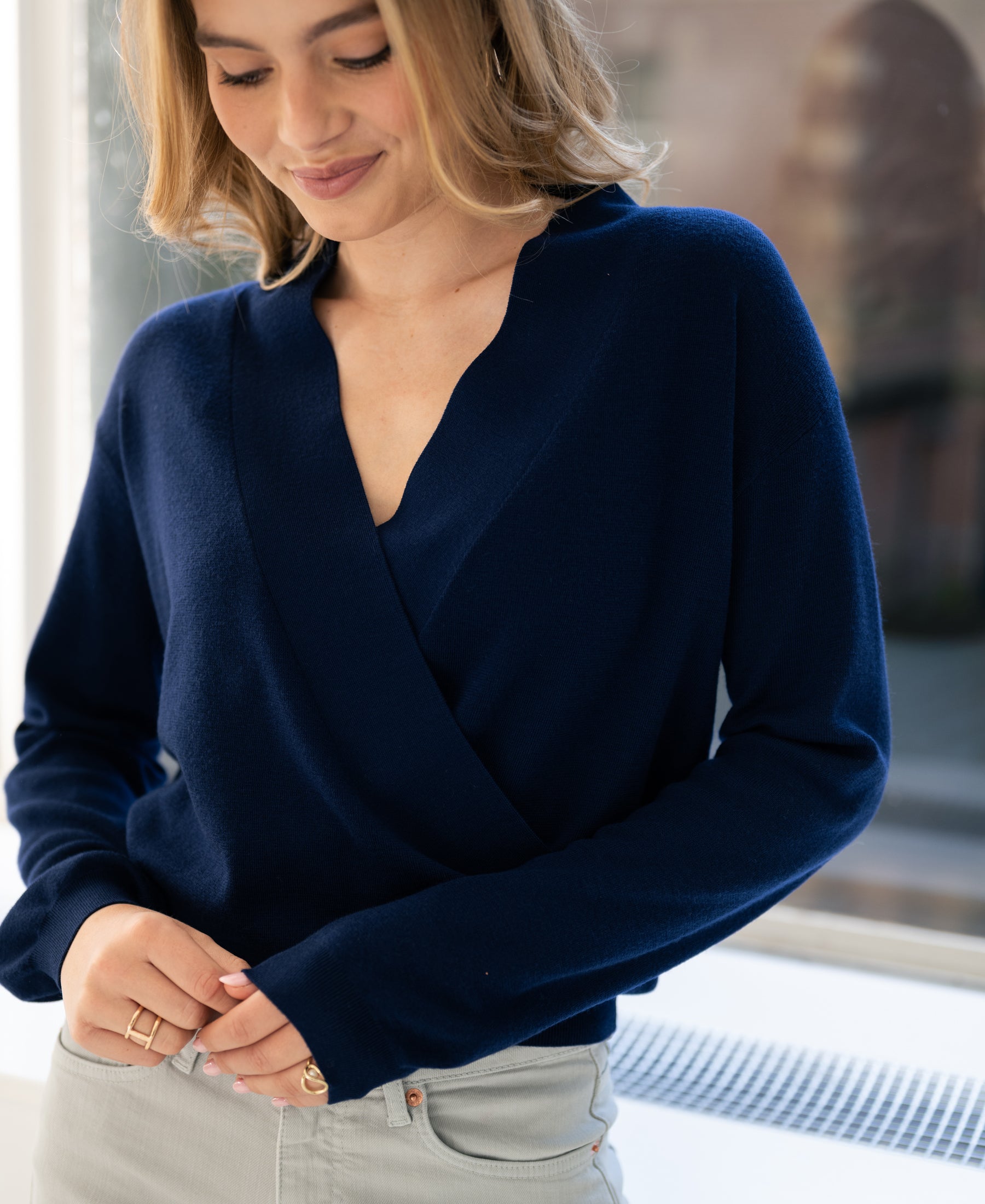 Navy LES CINQ wrap sweater by PLEIN PUBLIQUE, shown on a smiling woman by the window.