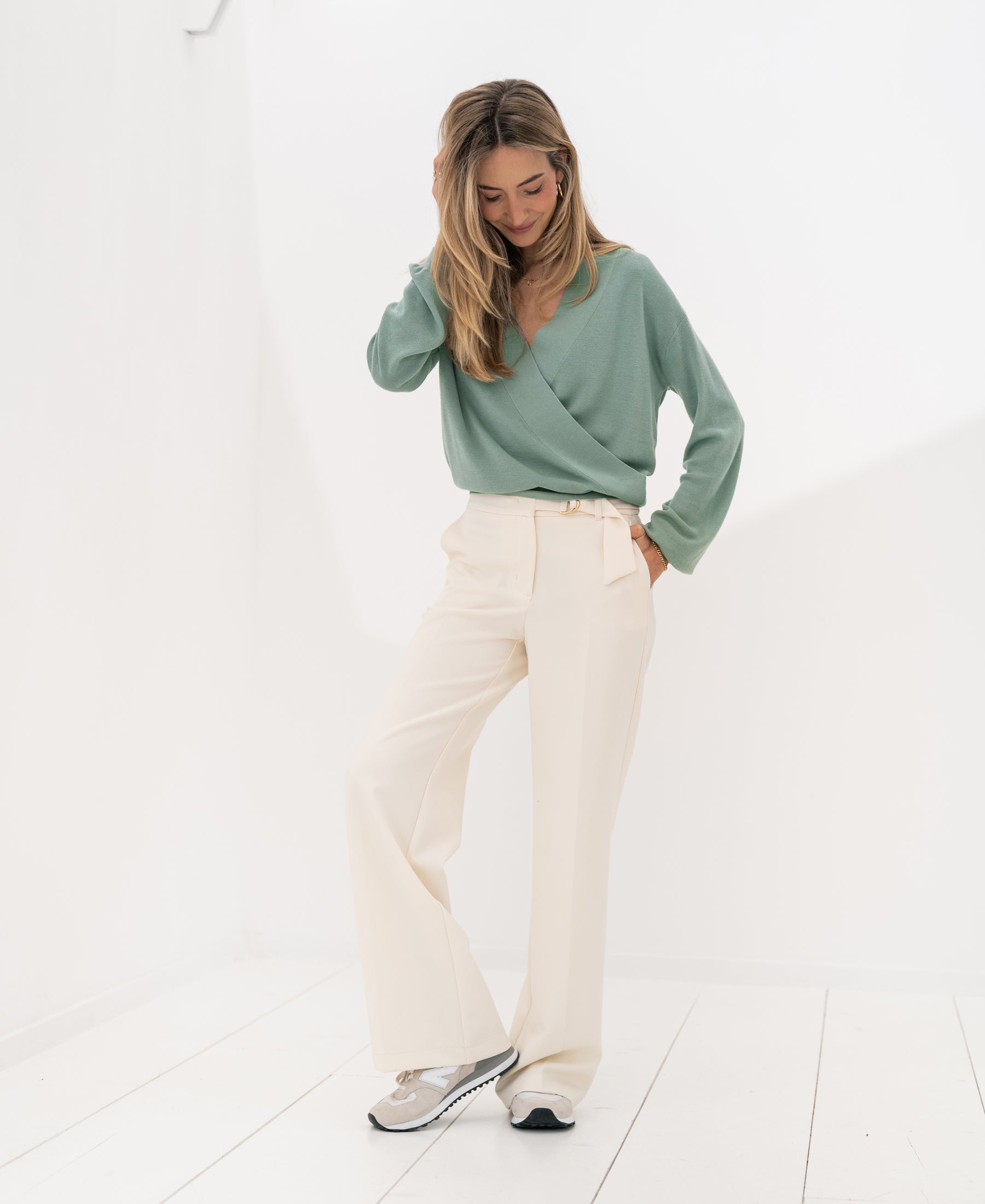Woman wearing cream LA WENDE trousers stands in a bright room, smiling and touching her hair.