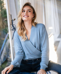 Woman wearing a blue PLEIN PUBLIQUE wrap sweater (LES CINQ) sits by a window.