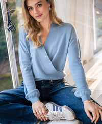 Woman wearing a PLEIN PUBLIQUE wrap sweater LES CINQ in jeans blue sits by a window, facing the camera.