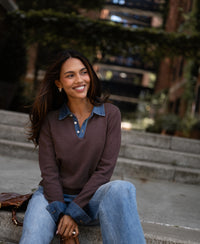 Woman sitting on outdoor steps, smiling in a brown cotton sweater-blouse with greenery in the background.