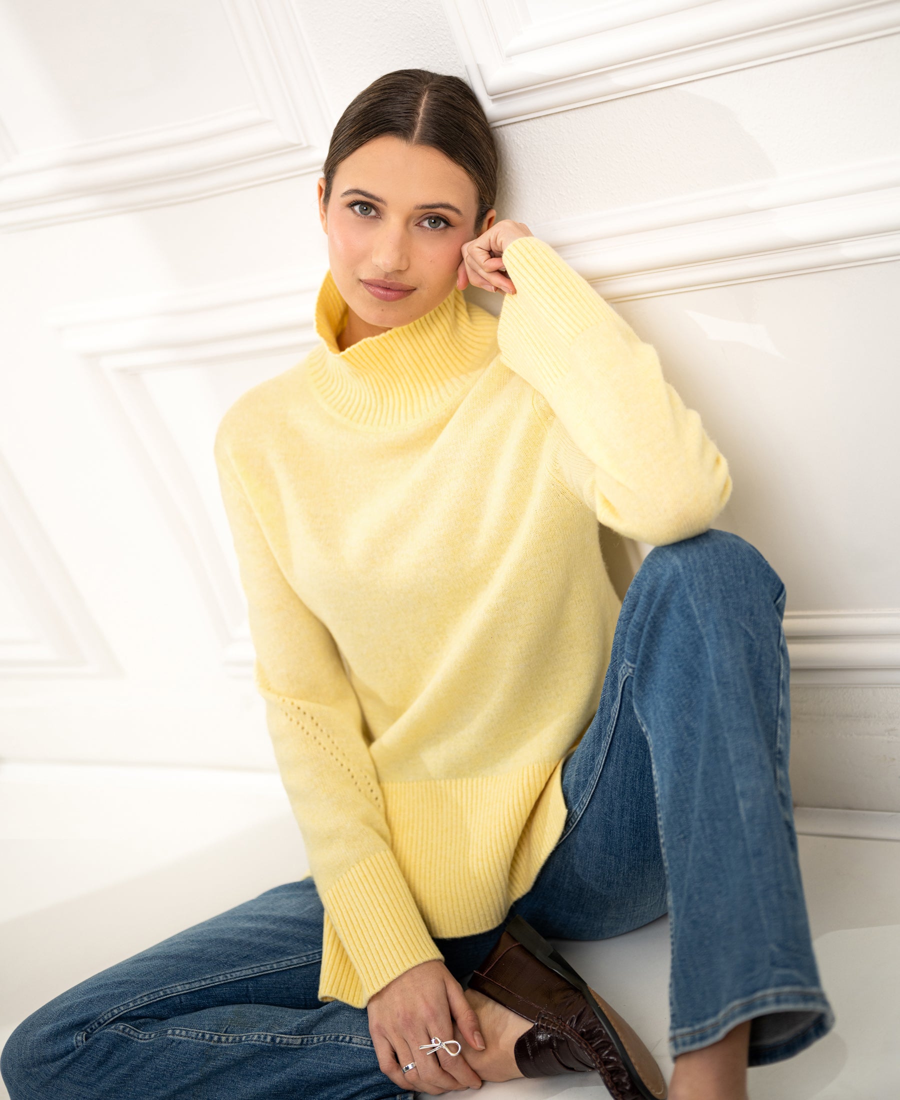 Woman wearing a PLEIN PUBLIQUE LA COZY turtleneck sweater in light yellow sits on the floor, leaning against a white wall.