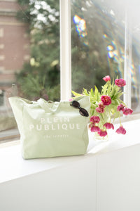 A green canvas tote bag by PLEIN PUBLIQUE sits on a windowsill with pink tulips.