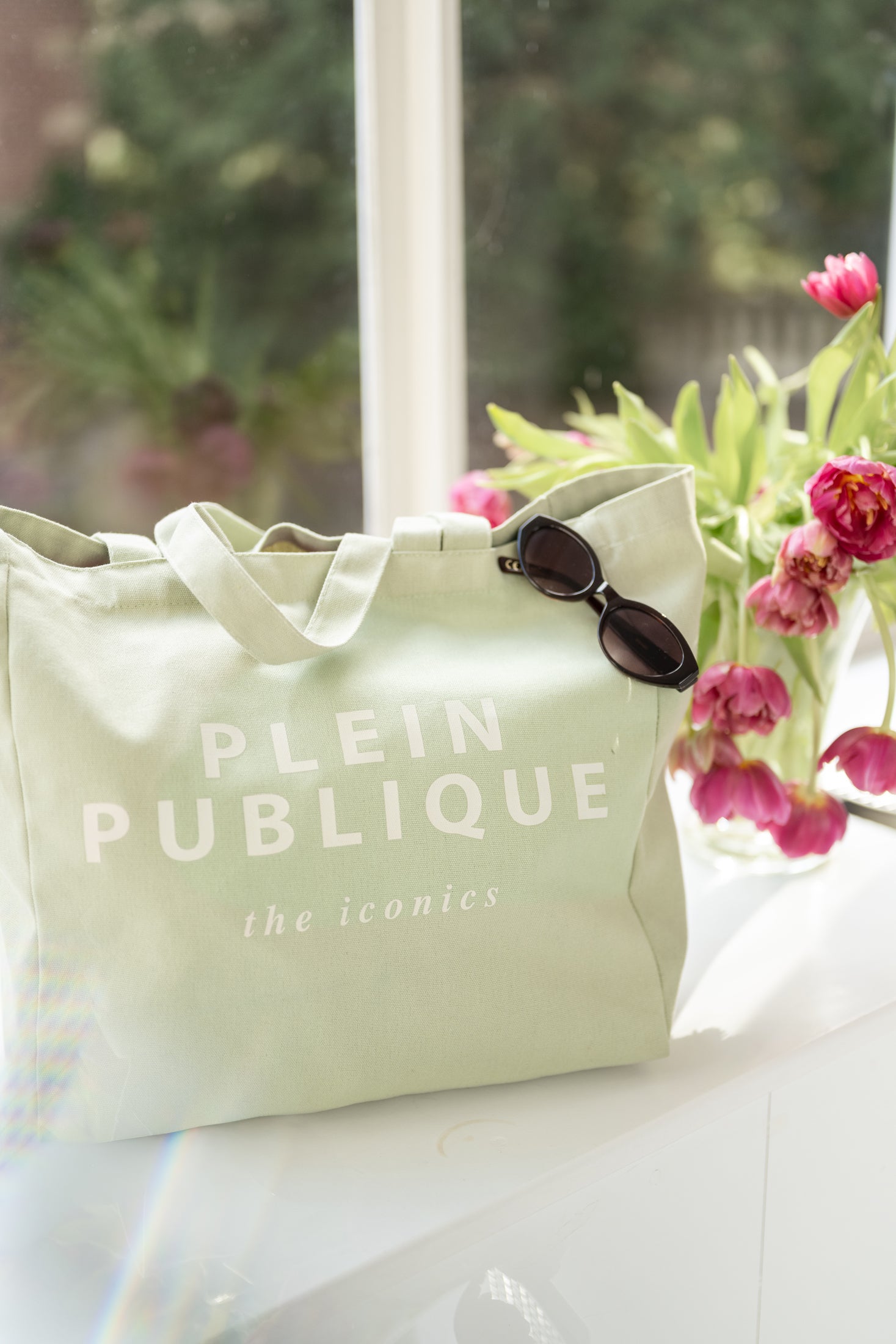 A trendy green canvas tote bag by PLEIN PUBLIQUE is placed on a sunny windowsill.