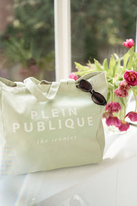 A trendy green canvas tote bag by PLEIN PUBLIQUE is placed on a sunny windowsill.