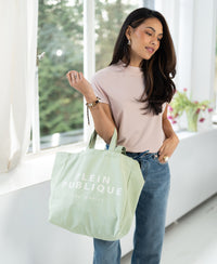 PLEIN PUBLIQUE green canvas tote bag, shown being held indoors by a window.