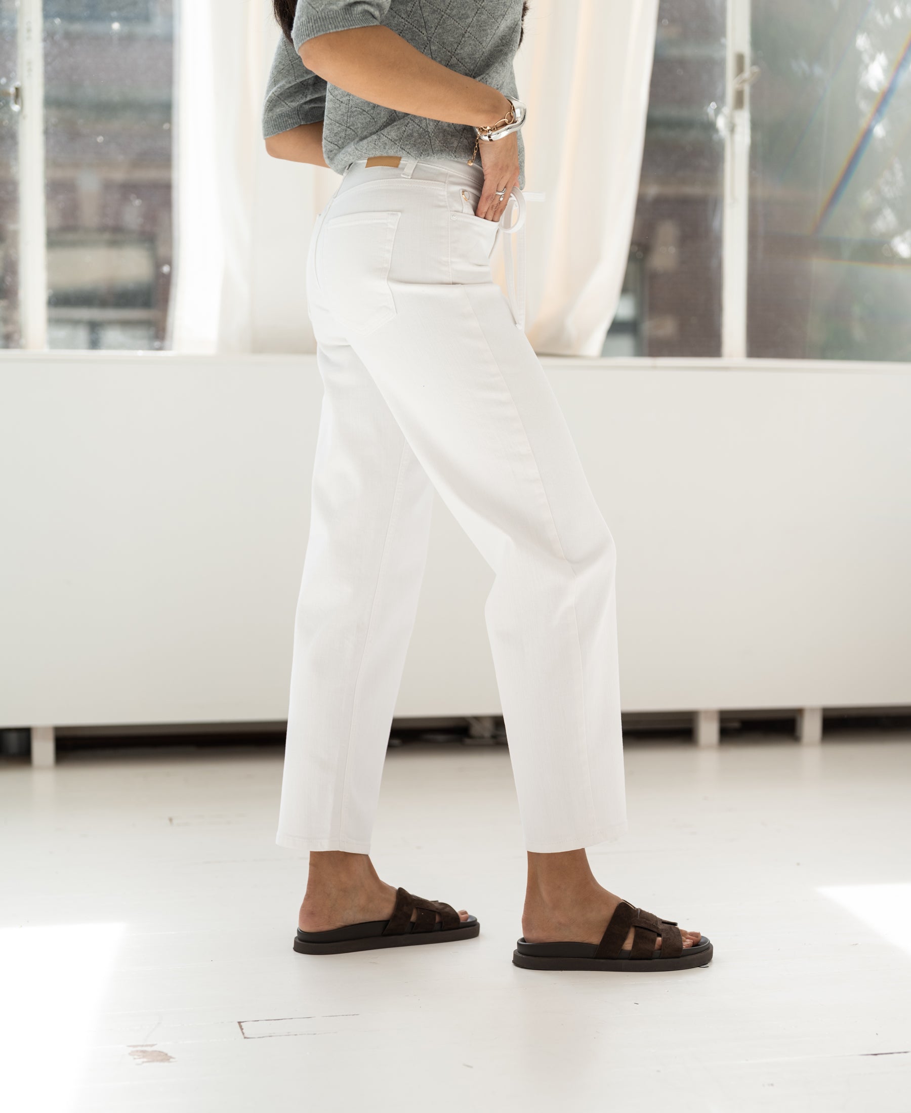 Person wears white cropped jeans, standing by a window indoors.
