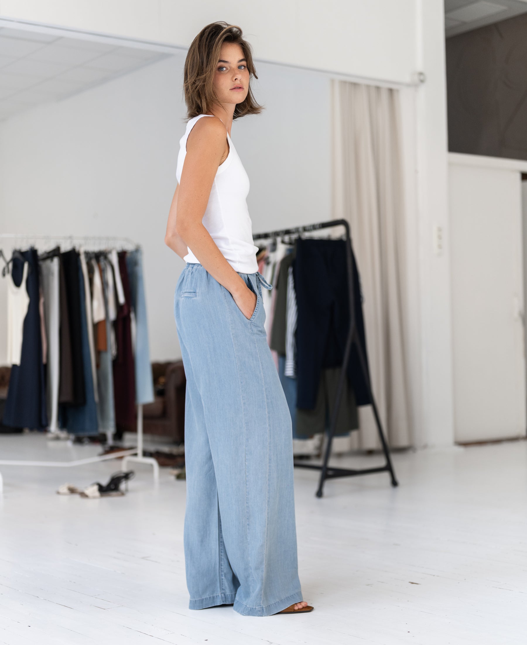 Woman wearing light-wash Tencel denim jeans stands by racks in a bright room.