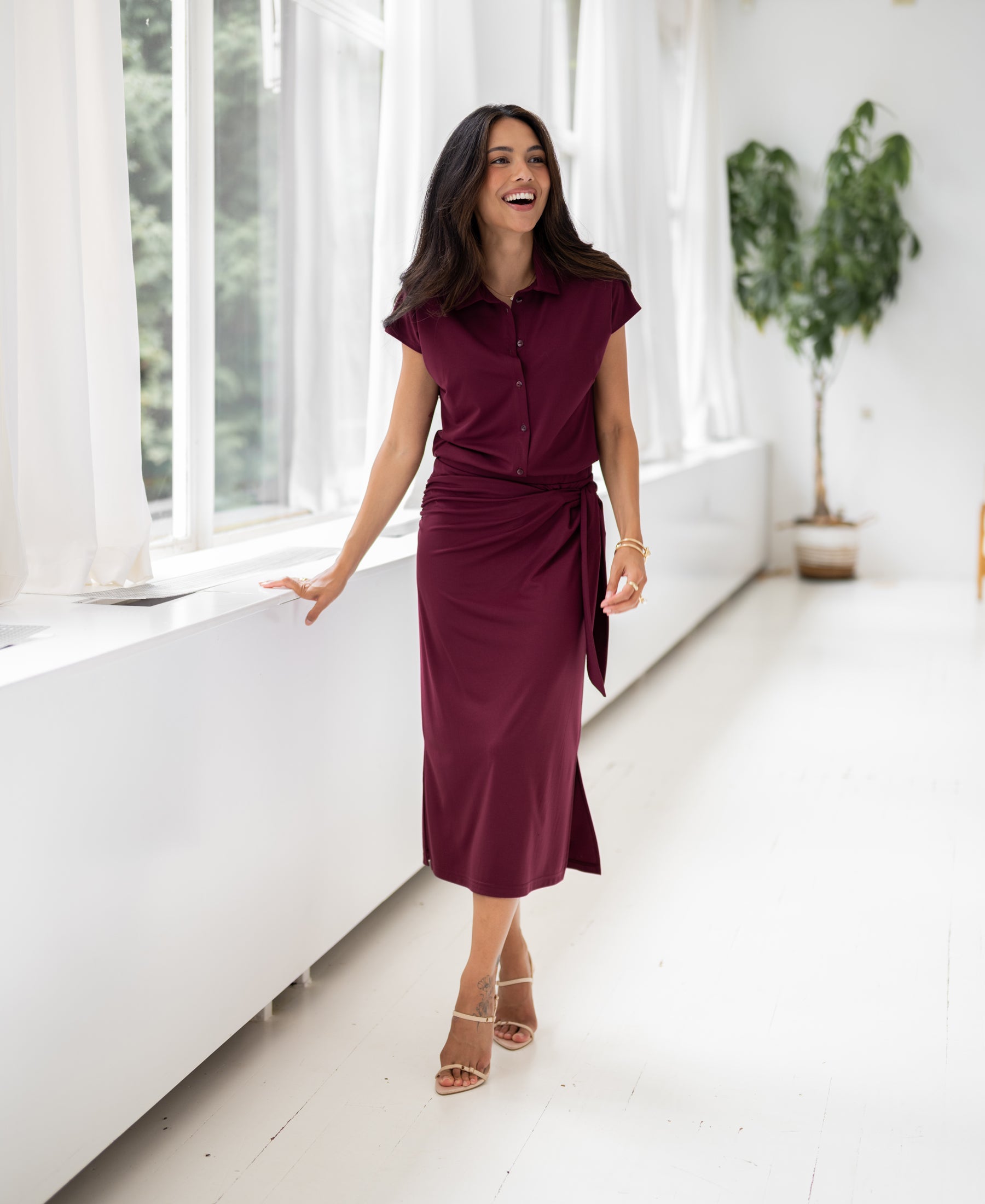 Woman wearing the Plein Publique wine-colored travel dress stands indoors near large windows and a plant.