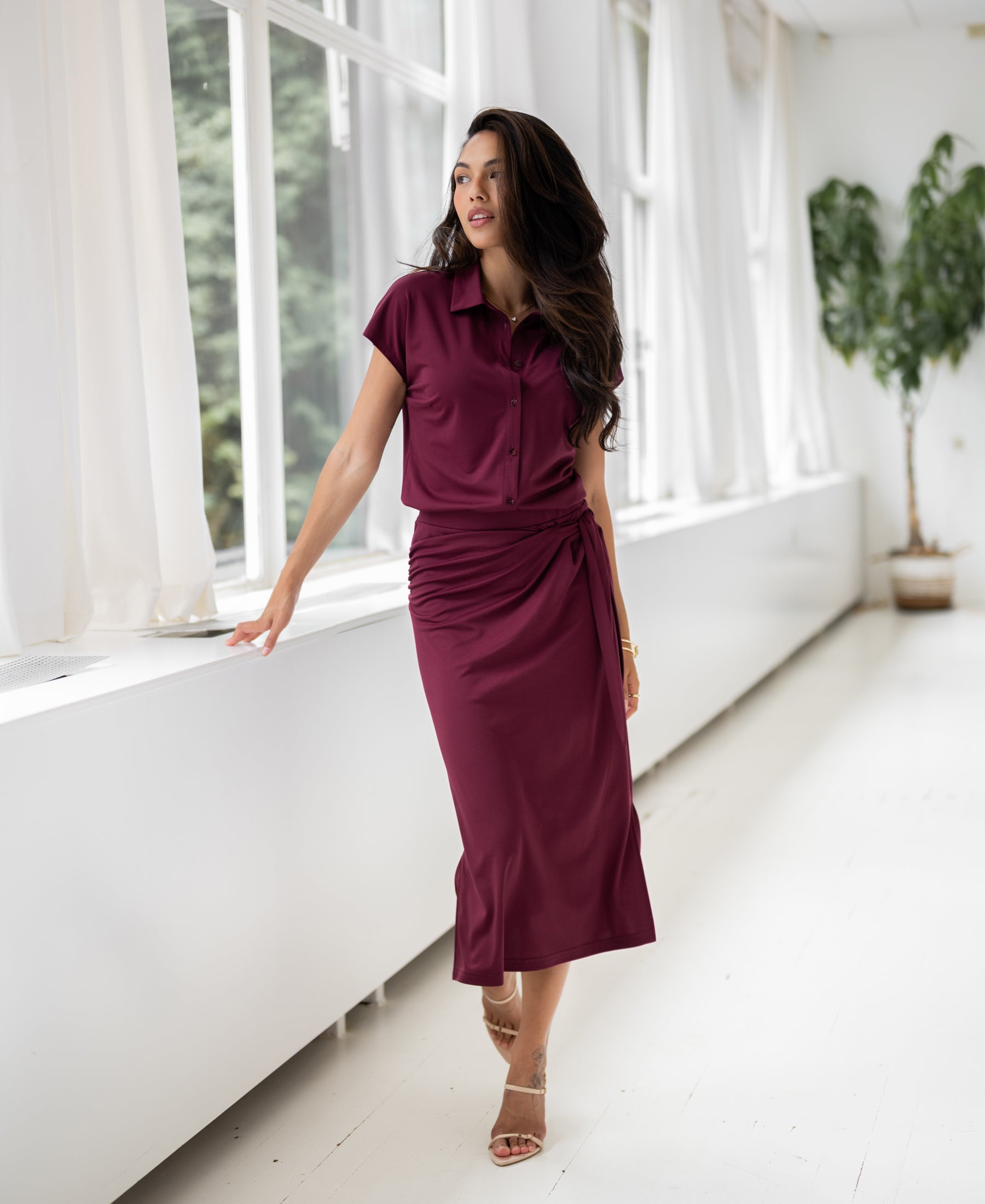 A woman wears a Plein Publique wine-colored travel blouse while standing by large windows in a bright, airy room.