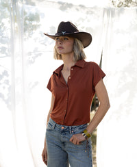 Woman outdoors by sheer white curtains wears the PLEIN PUBLIQUE Terra brown travel blouse.