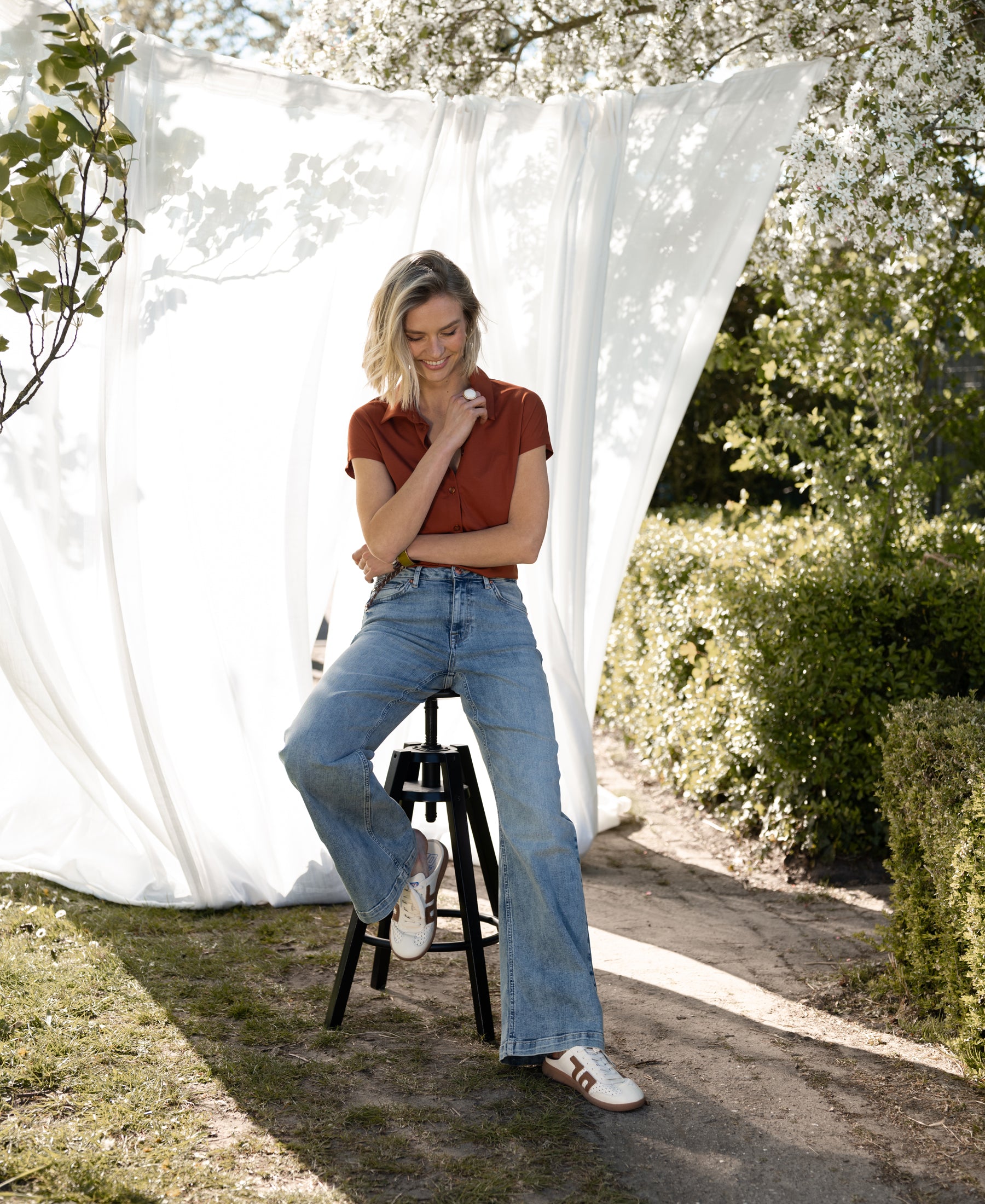 Terra-colored LA DELON Travelblouse by PLEIN PUBLIQUE displayed outdoors among white curtains and greenery.
