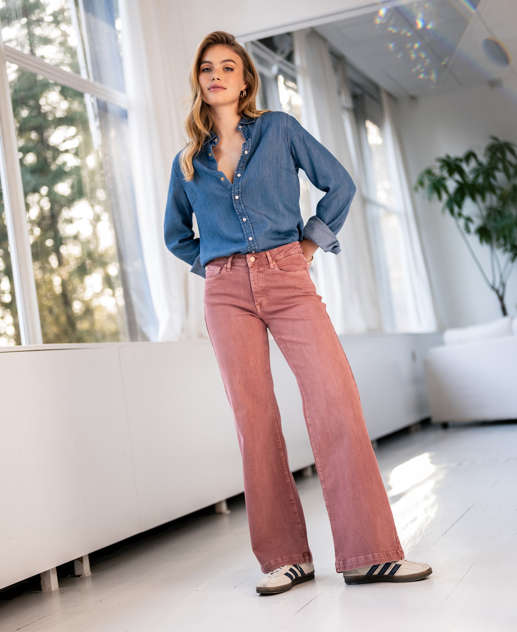 Woman standing indoors in sunlight, wearing pink LES DEUX jeans by PLEIN PUBLIQUE.