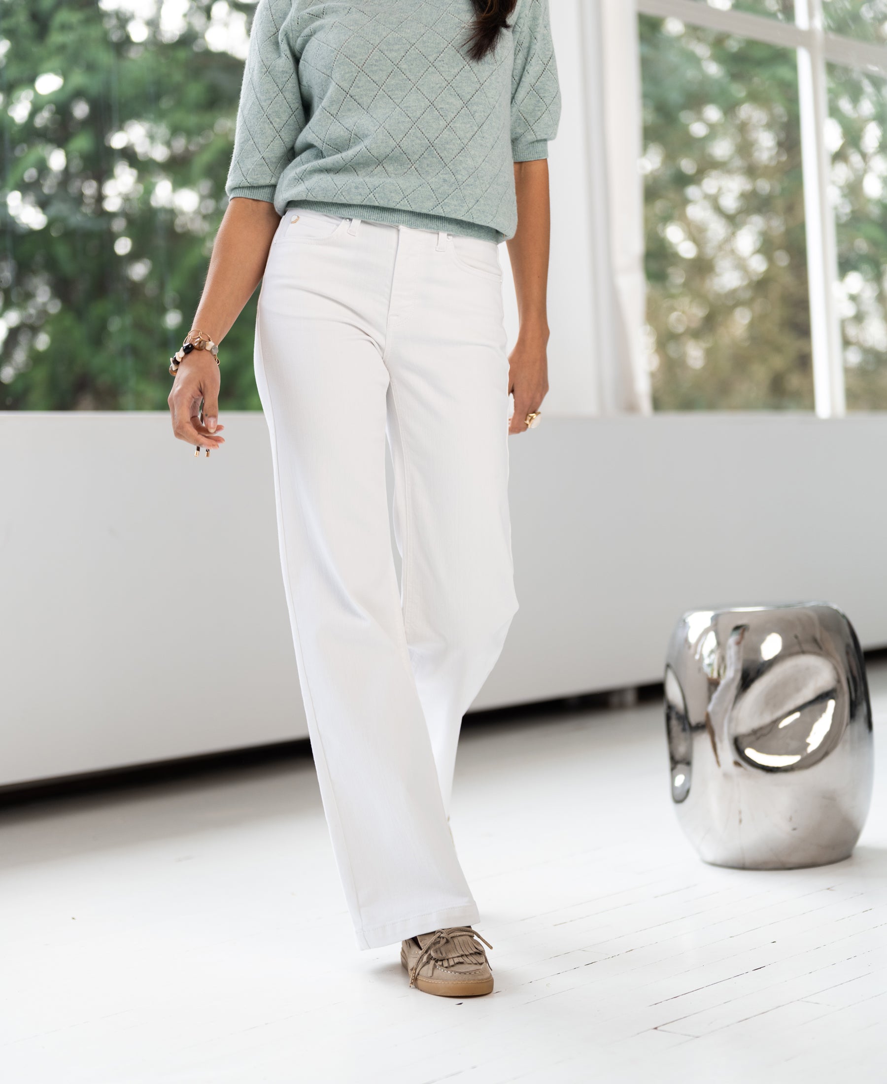 Wide leg jeans in light denim, shown indoors near a silver stool with large windows in the background.