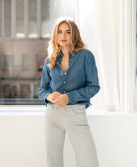 Tencel denim blouse in stonewash blue, shown worn by a woman standing by a sunlit window.