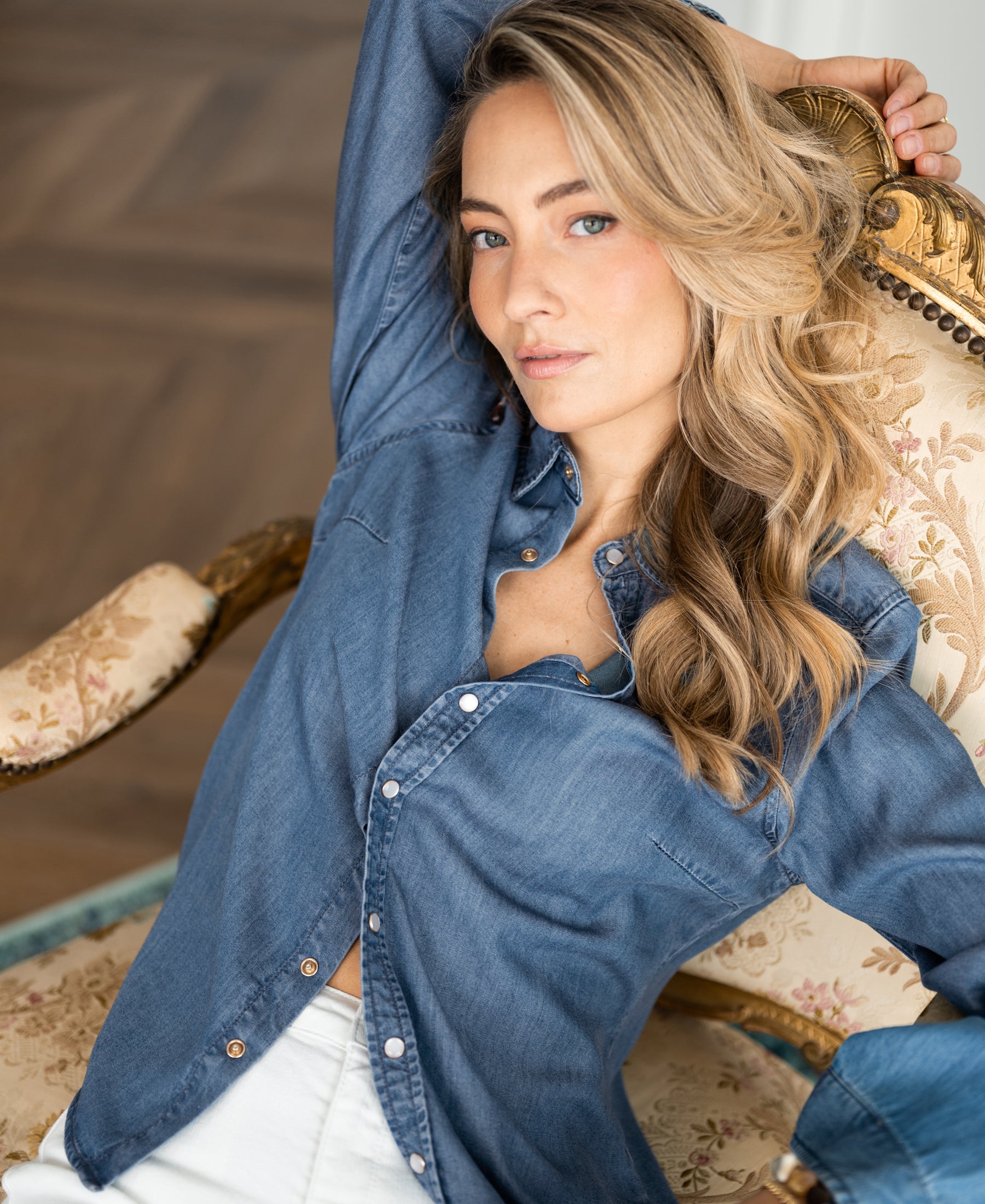 Woman wearing a stonewash Tencel denim blouse, seated with one arm raised.