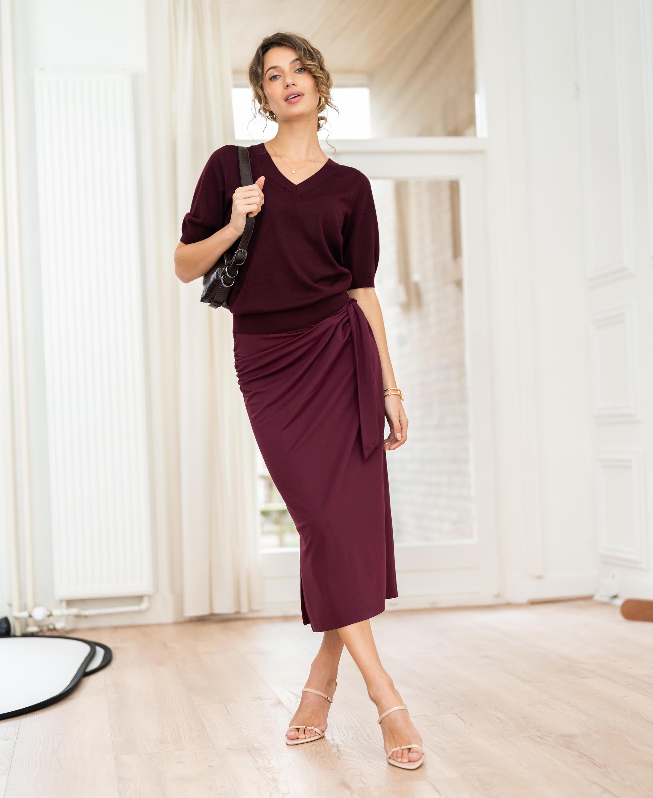 Woman wearing a burgundy PLEIN PUBLIQUE LE MAEL travel skirt poses indoors on a wooden floor.