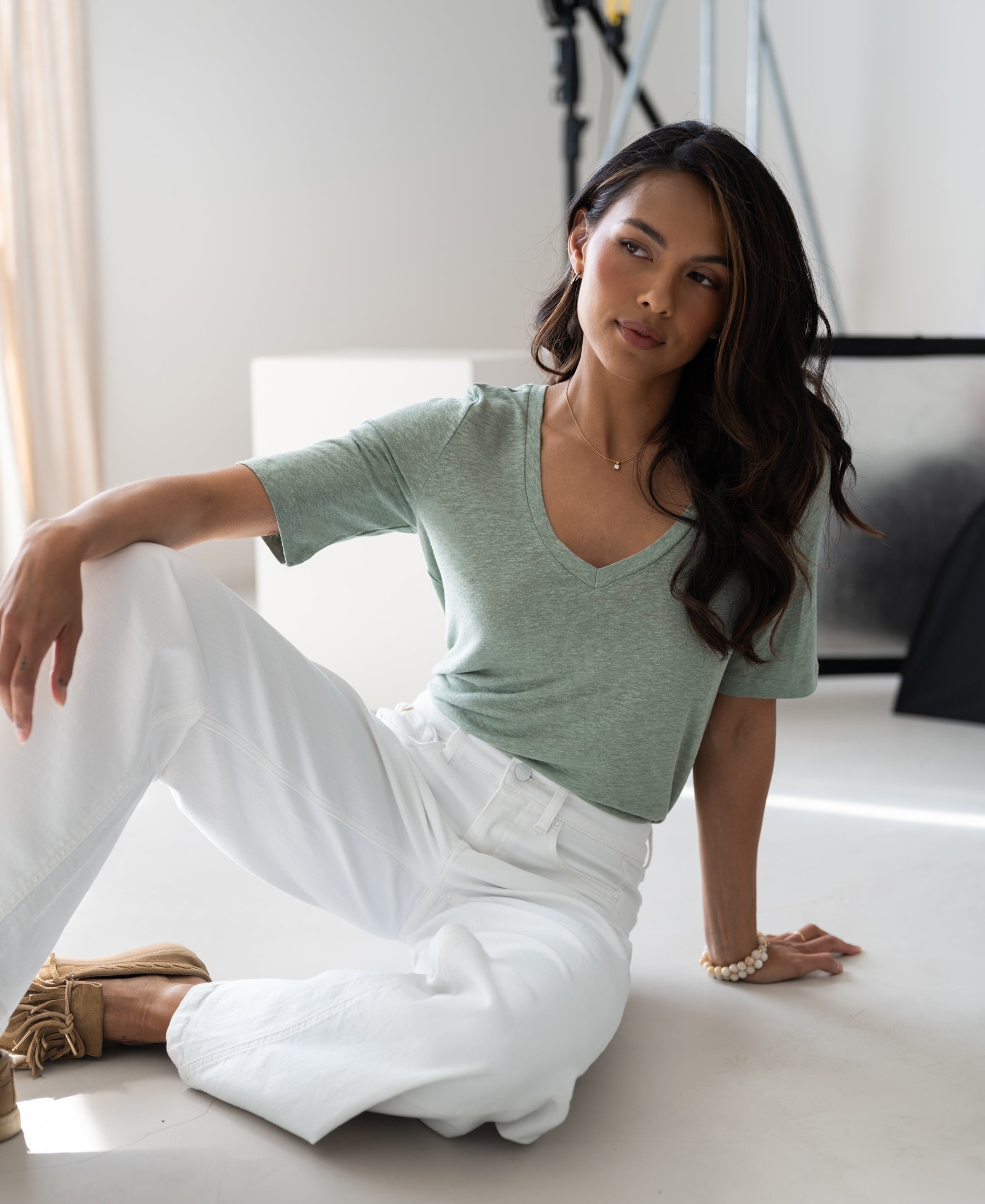 Woman wears light mist green linen T-shirt by PLEIN PUBLIQUE, sitting in a bright modern studio.