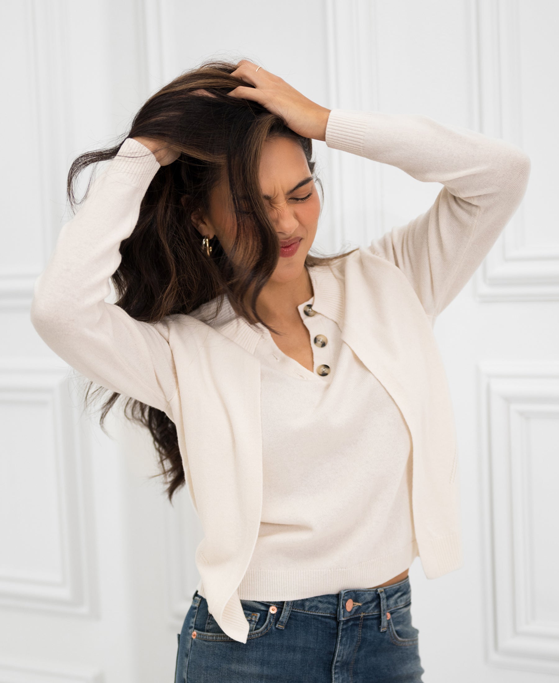 Woman wears ivory knit sweater with matching cardigan indoors, hands in hair, looking frustrated.