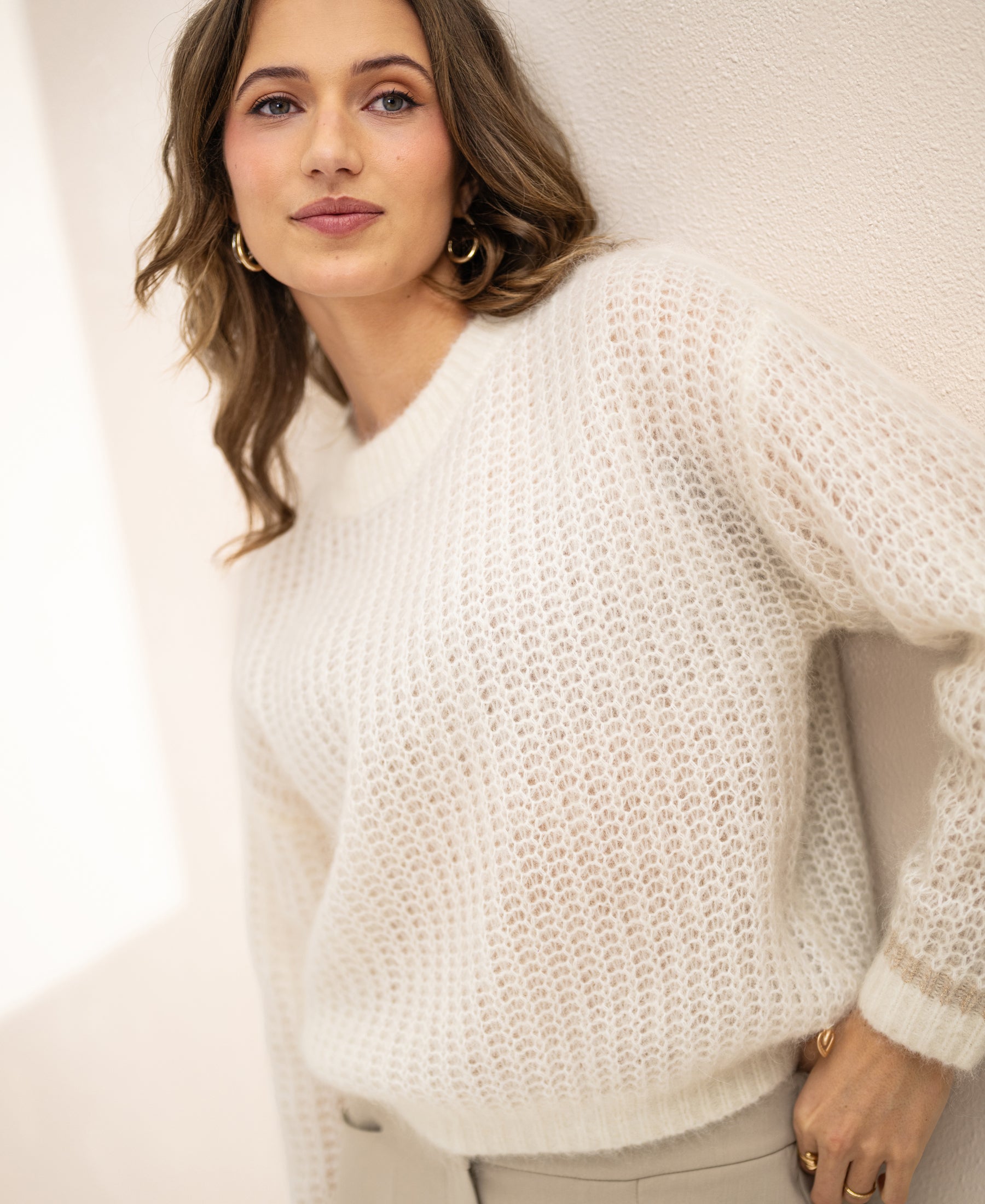 Woman wearing PLEIN PUBLIQUE's LA DOUCE ivory white mohair sweater stands by a light wall, looking at the camera.