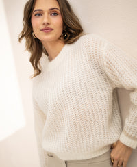 Woman wearing PLEIN PUBLIQUE's LA DOUCE ivory white mohair sweater stands by a light wall, looking at the camera.
