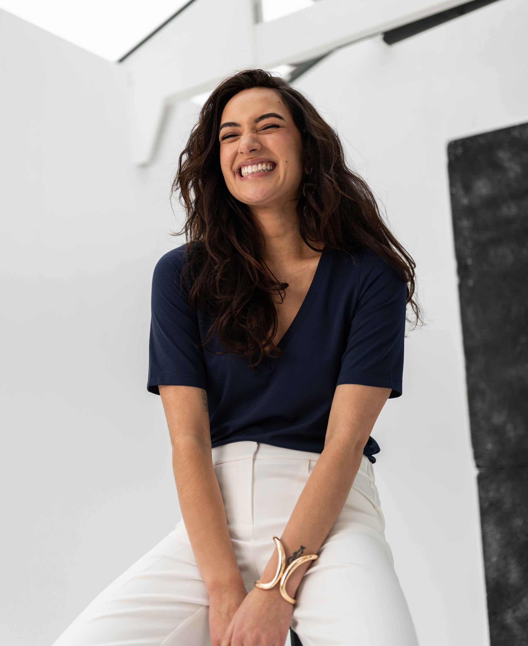Woman wearing a PLEIN PUBLIQUE V-neck T-shirt LA VERONIQUE in dark blue, seated in a modern space.