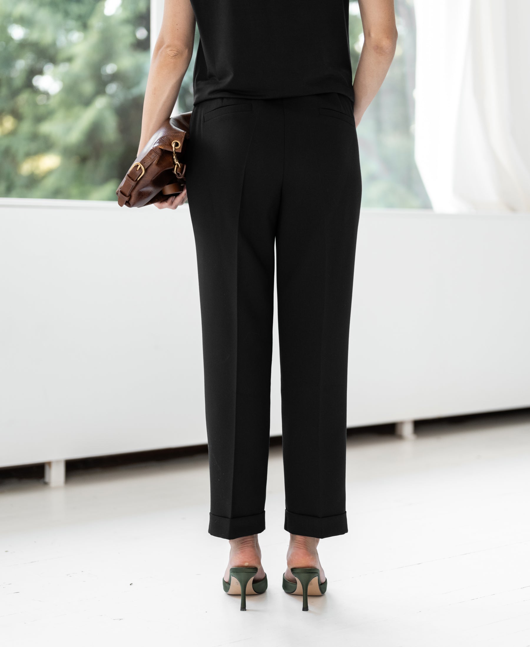 Woman wearing PLEIN PUBLIQUE 7/8 FAUBOURG black pants standing by a window with greenery outside.