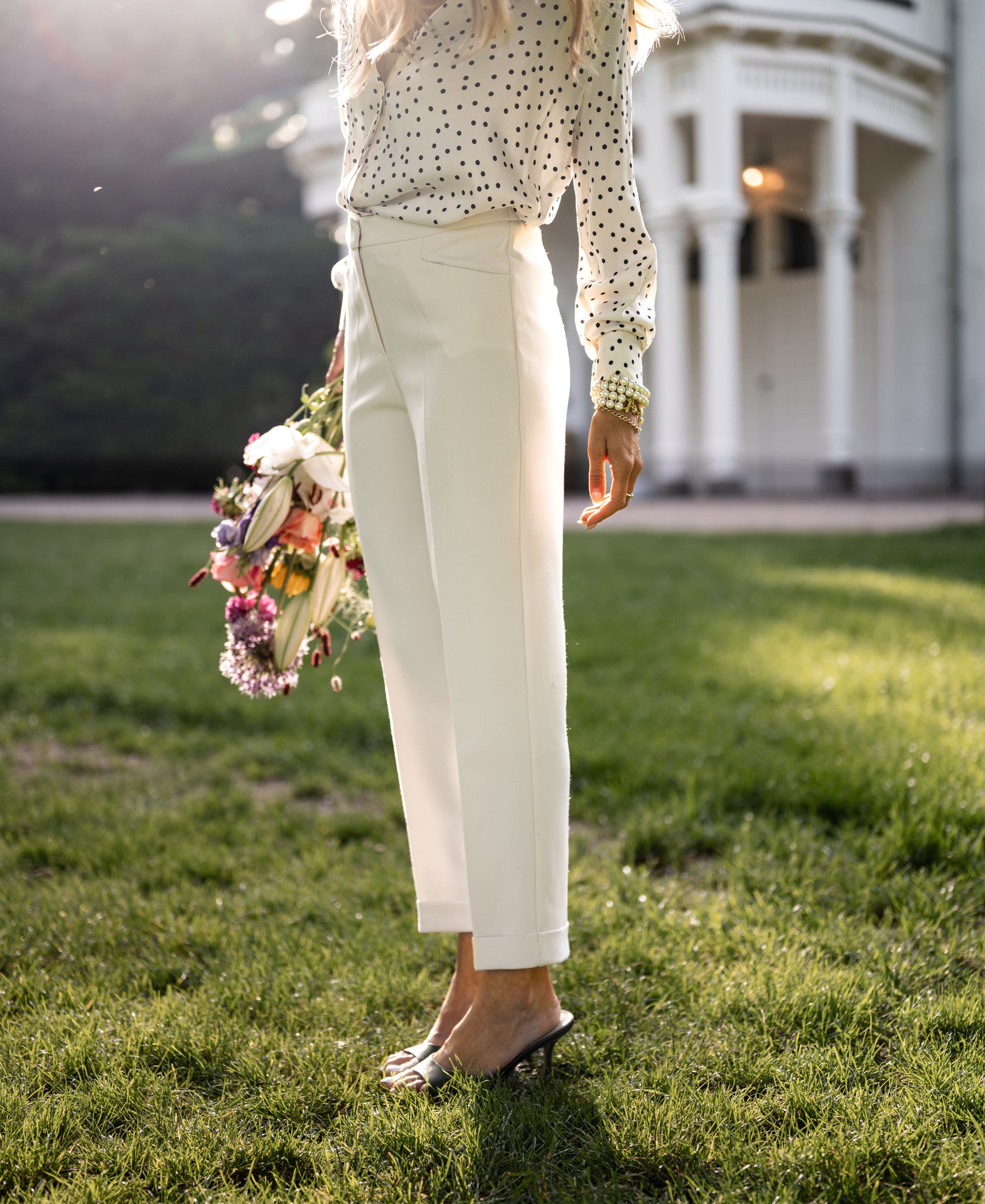 Woman wearing cream tapered pants stands on grass near a white building.