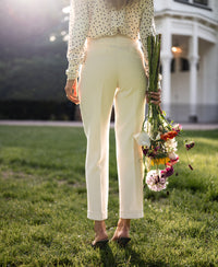 Woman wearing cream tapered pants, standing on grass near a white building.