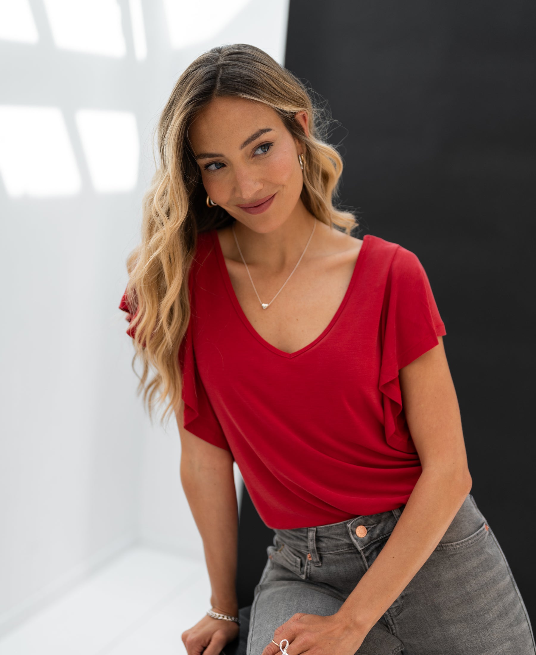 Woman wearing a red Plein Publique Twoway LA FIORA top sits in a bright, black-and-white room.