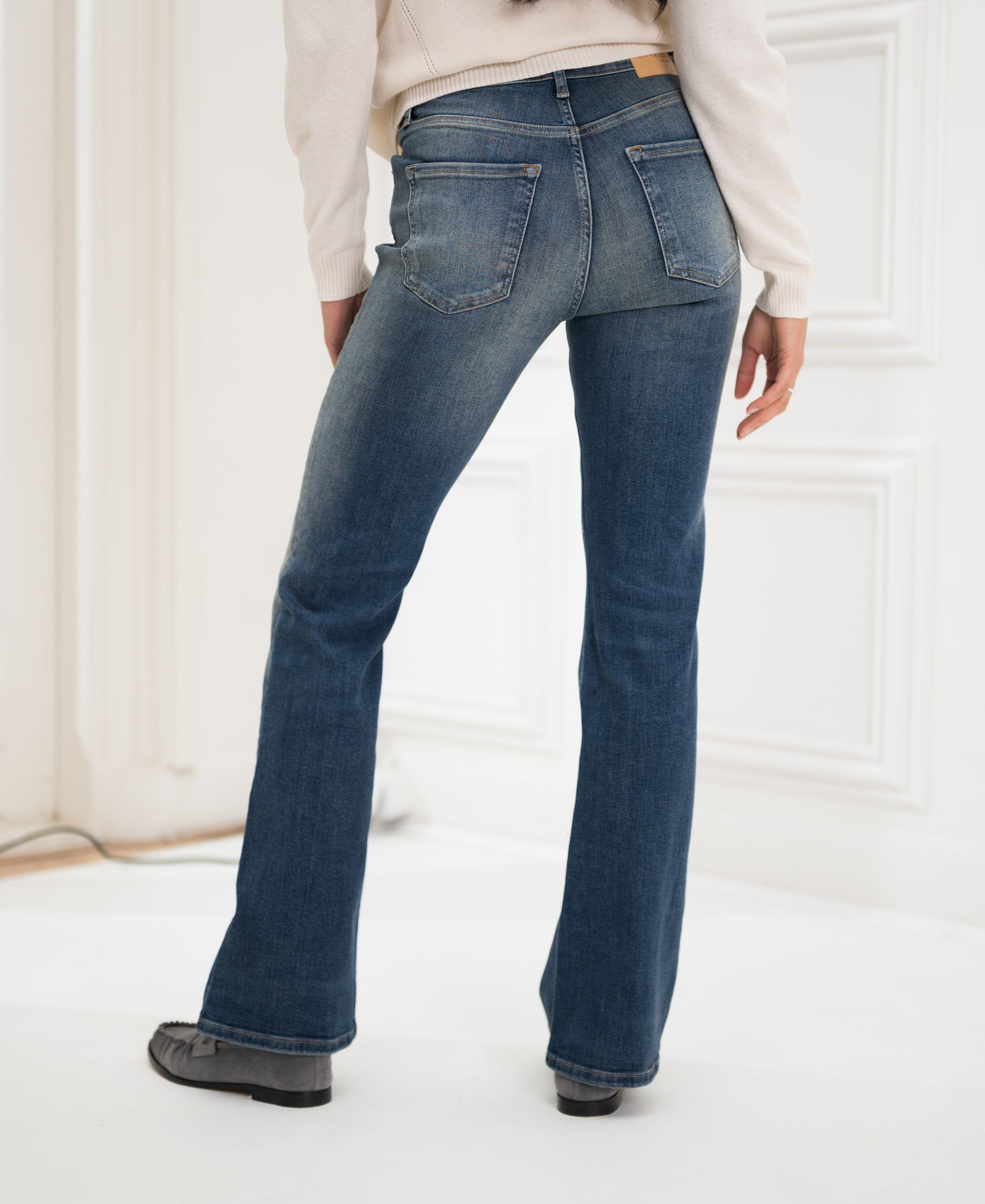 Person wearing blue vintage flared jeans by PLEIN PUBLIQUE, seen from behind indoors.