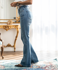 Light stone wash flared jeans in a full-length indoor view, shown by a mirror and table with sunlight streaming in.