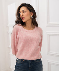 Woman wears pink PLEIN PUBLIQUE LA FRAMBOISE sweater, standing indoors by a white paneled wall.