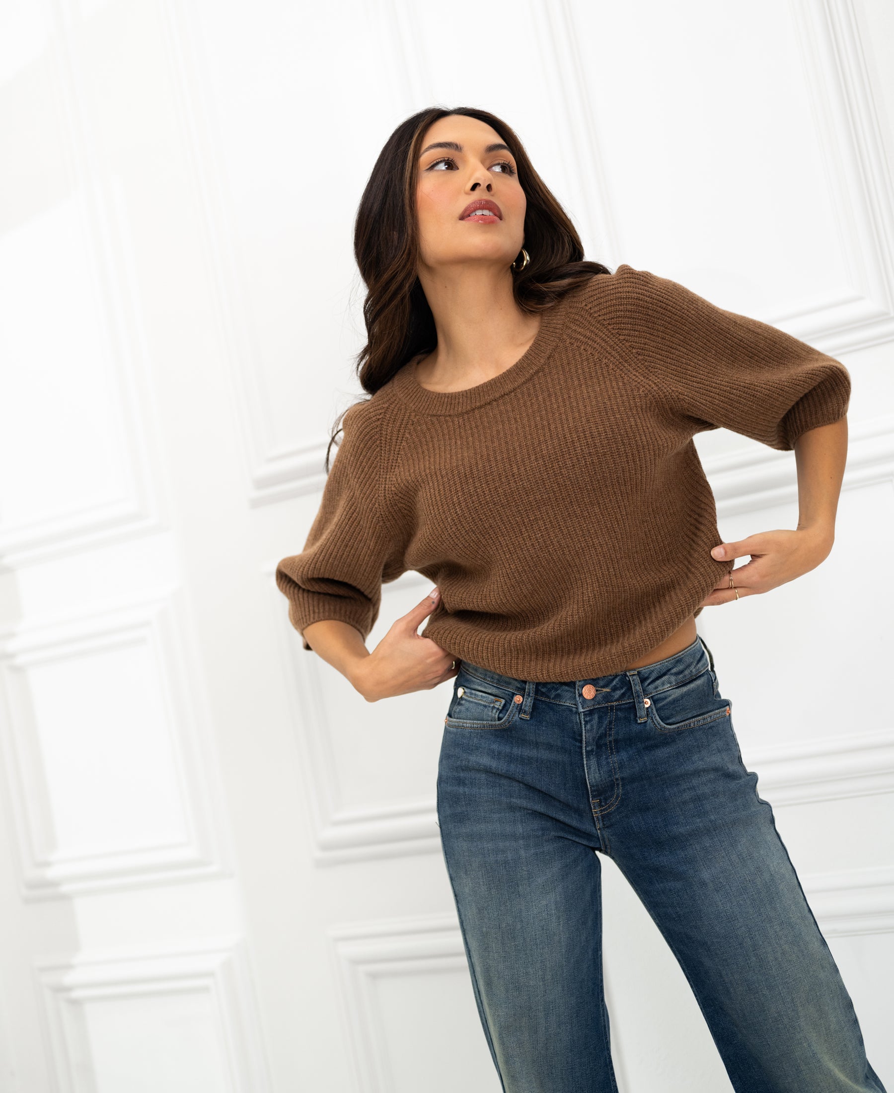 Woman wearing a tobacco-colored LA FRAMBOISE sweater by PLEIN PUBLIQUE stands against a white paneled wall.