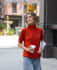 Woman wearing a cayenne short sleeve turtleneck smiles on the sidewalk.