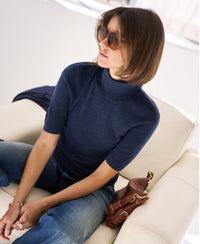 Short sleeve turtleneck sweater in blue, shown seated on a white chair.