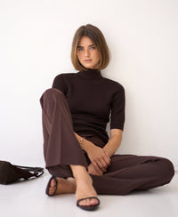 Brown short-sleeved turtleneck sweater, Pure Choco color, shown on a woman seated indoors against a white wall.