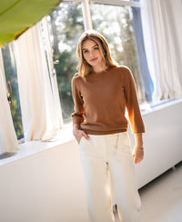 Woman wearing the LE FREJUS Merino sweater by PLEIN PUBLIQUE poses by a sunny window, looking at the camera.