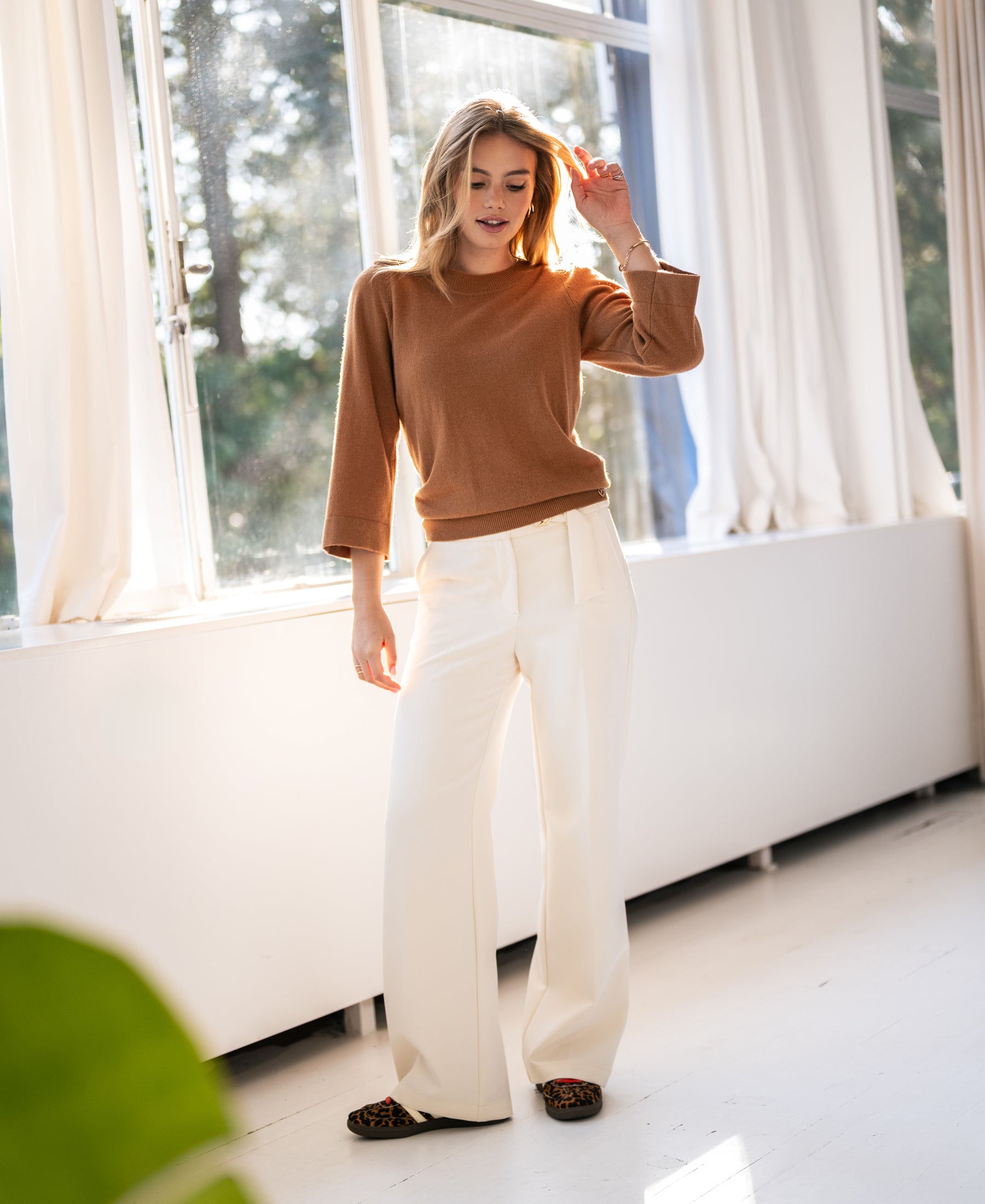 A woman stands in a sunlit room wearing the PLEIN PUBLIQUE Camel merino wool sweater.