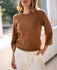 Camel merino wool sweater in a classic style, shown indoors near a window, hand in pocket.