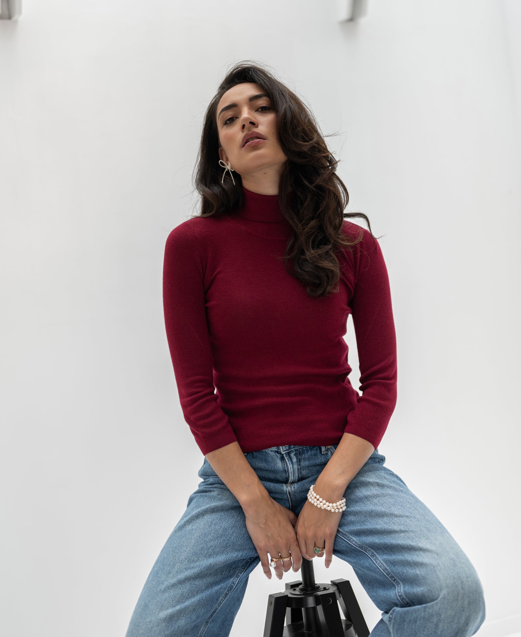 Woman wearing a PLEIN PUBLIQUE LE FABIAN dark red turtleneck sweater, seated on a stool.