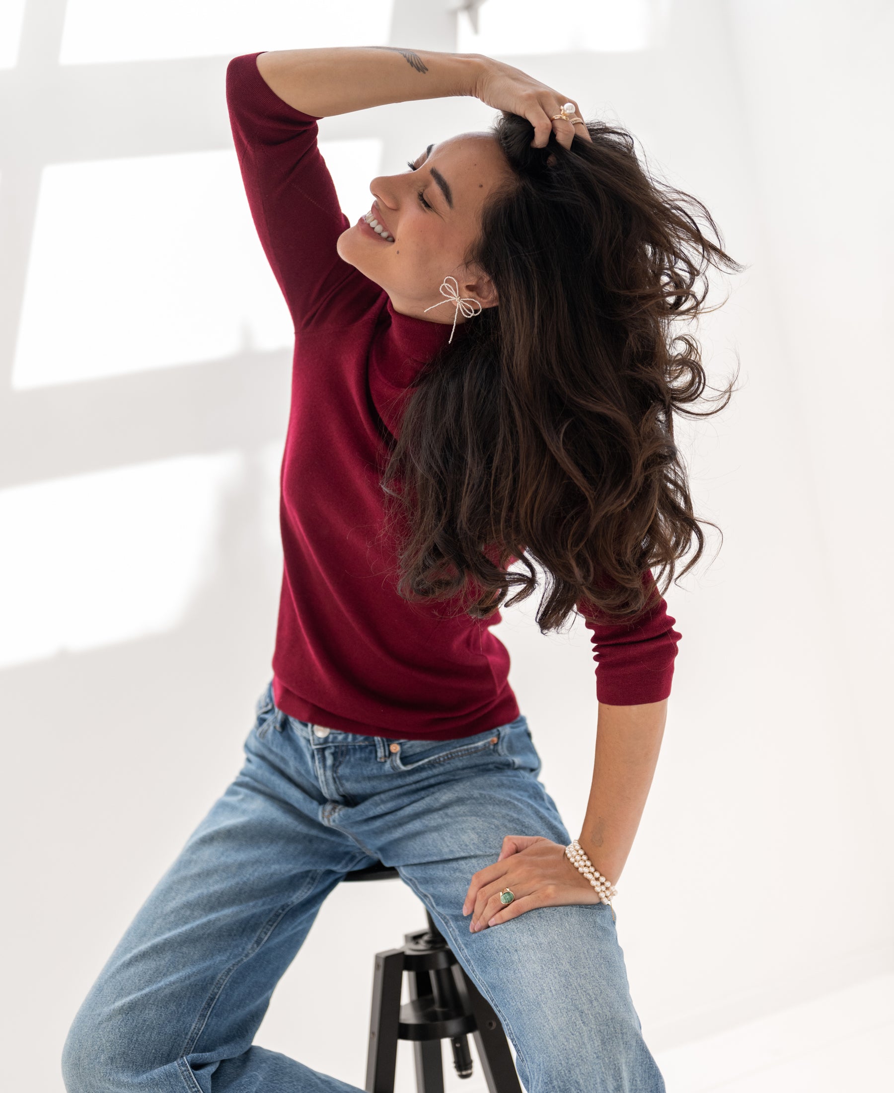 Woman wearing a PLEIN PUBLIQUE LE FABIAN dark red turtleneck sweater, shown seated in bright natural light.