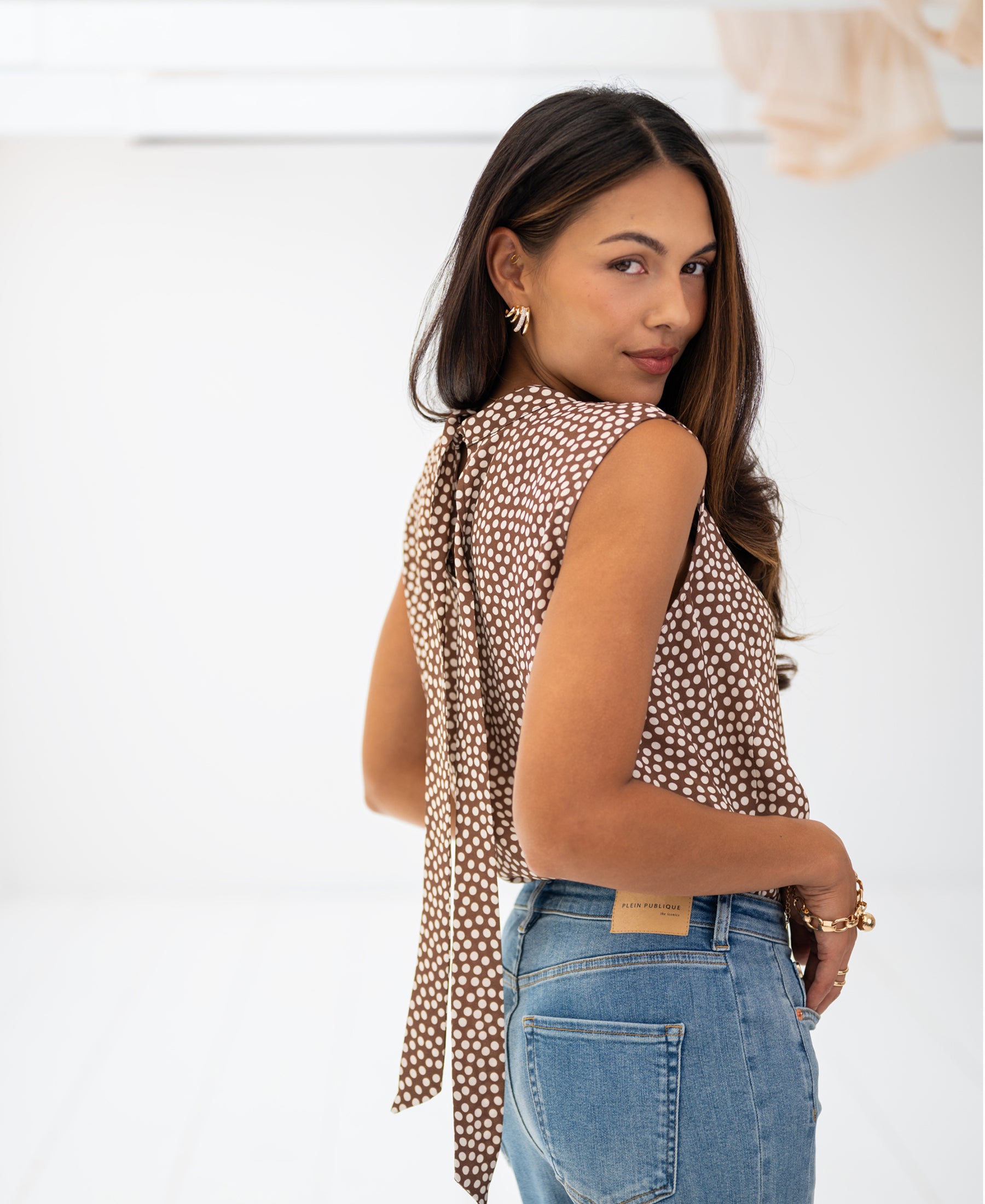 Woman wearing a mocha mousse-cream polka dot top looks over her shoulder in a bright room.