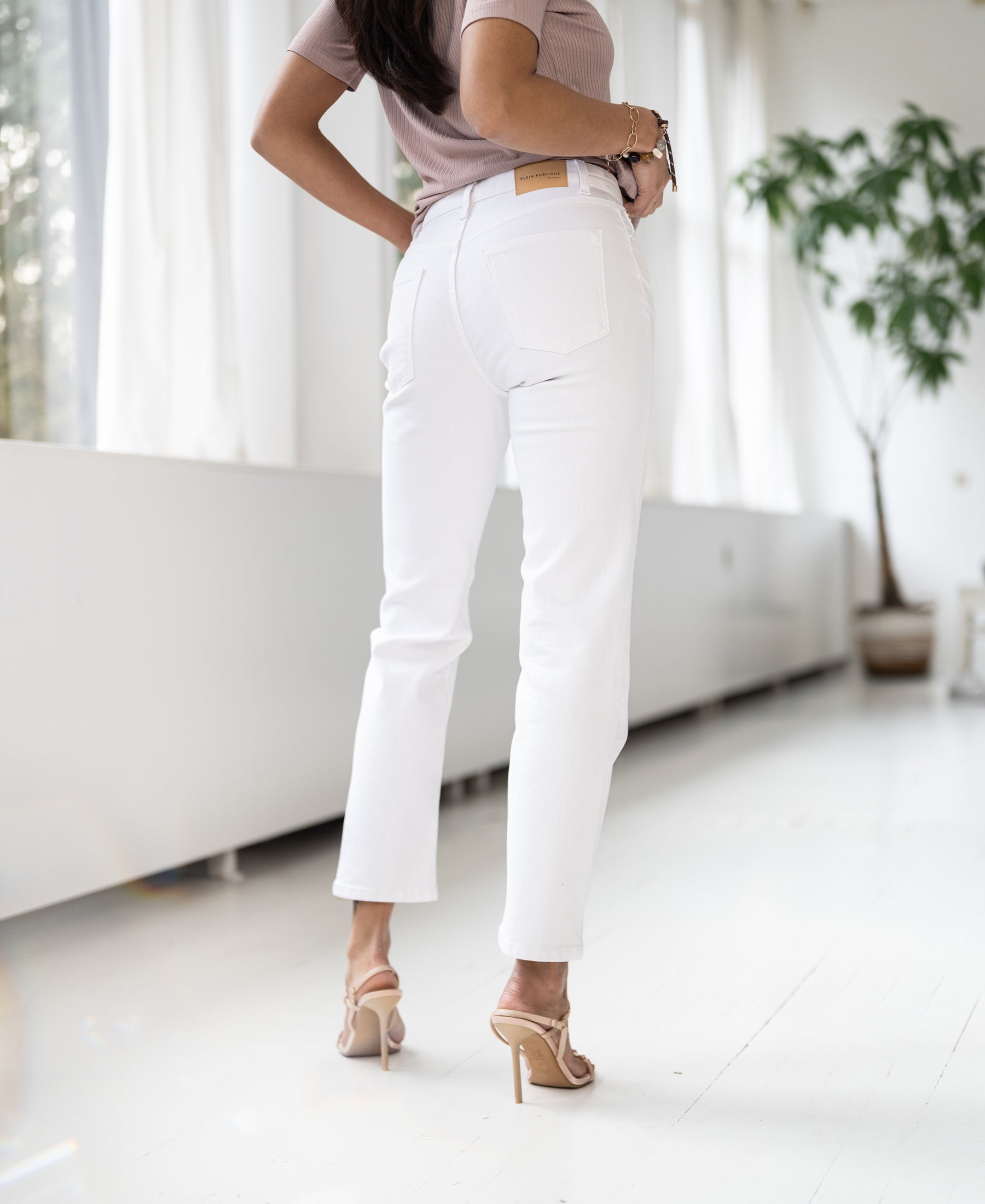 Back view of woman wearing cropped jeans in light color, standing indoors with natural light streaming through large windows.